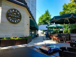 An outdoor café setting with neatly arranged tables and chairs under green umbrellas. A sign on the wall reads 'Café in the Square Florya' adorned with lights. Decorative flower boxes with green and red plants line the pathway, which is paved with light-colored tiles. The background shows blurred figures walking and a lush green landscape.