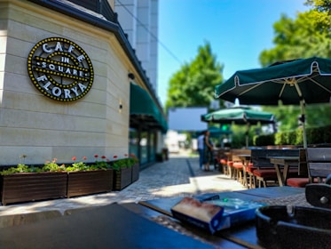 An outdoor café setting with neatly arranged tables and chairs under green umbrellas. A sign on the wall reads 'Café in the Square Florya' adorned with lights. Decorative flower boxes with green and red plants line the pathway, which is paved with light-colored tiles. The background shows blurred figures walking and a lush green landscape.