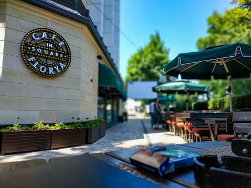 An outdoor café setting with neatly arranged tables and chairs under green umbrellas. A sign on the wall reads 'Café in the Square Florya' adorned with lights. Decorative flower boxes with green and red plants line the pathway, which is paved with light-colored tiles. The background shows blurred figures walking and a lush green landscape.