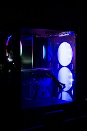 A computer case with visible components illuminated by blue and purple LED lights. The interior features a large fan, cables, and various hardware elements, including heat sinks and circuit boards. The lighting creates a futuristic and tech-focused atmosphere.