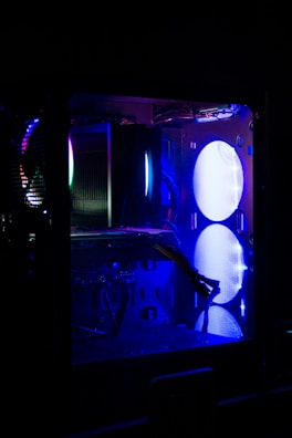 A computer case with visible components illuminated by blue and purple LED lights. The interior features a large fan, cables, and various hardware elements, including heat sinks and circuit boards. The lighting creates a futuristic and tech-focused atmosphere.