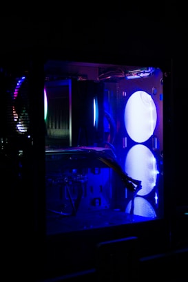 A computer case with visible components illuminated by blue and purple LED lights. The interior features a large fan, cables, and various hardware elements, including heat sinks and circuit boards. The lighting creates a futuristic and tech-focused atmosphere.