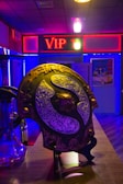 A room with dim lighting features a prominently displayed decorative shield with ornate metalwork on a countertop. Behind the shield, a neon sign with 'VIP' in red letters is visible. The room is filled with ambient lighting in shades of blue and purple, and there are additional items and posters visible in the background.