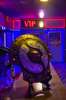 A room with dim lighting features a prominently displayed decorative shield with ornate metalwork on a countertop. Behind the shield, a neon sign with 'VIP' in red letters is visible. The room is filled with ambient lighting in shades of blue and purple, and there are additional items and posters visible in the background.