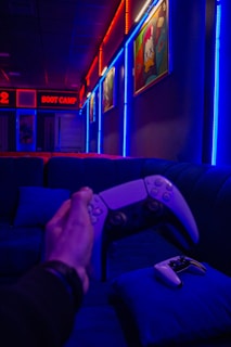 A person is holding a game controller in a dimly lit room with vibrant neon lights in red and blue. The room has comfortable seating along the walls adorned with colorful pop art or graffiti-style paintings. A second game controller rests on a blue pillow.