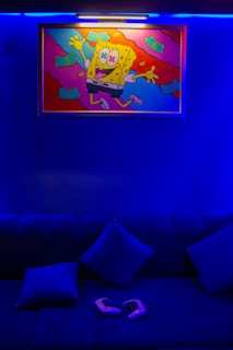 A vibrant painting featuring a cartoon character is mounted on a wall, illuminated by a light above. The character appears joyful, with cash floating around it. Below is a dark blue couch with two cushions, and a pair of gaming controllers placed on it. The room is bathed in a deep blue ambiance.