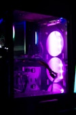 A close-up view of a computer case interior illuminated by purple and blue LED lights. Visible components include a graphics card, cables, and fans. The case has a modern, sleek design with transparent panels and vibrant lighting effects.