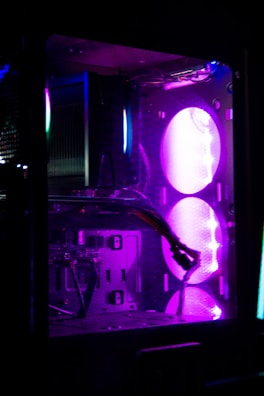 A close-up view of a computer case interior illuminated by purple and blue LED lights. Visible components include a graphics card, cables, and fans. The case has a modern, sleek design with transparent panels and vibrant lighting effects.
