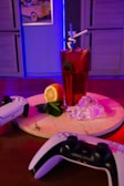 A tall glass of red beverage with a straw and mint leaves is placed on a round wooden board. Ice cubes and a half lemon are arranged next to the glass, which is brightly illuminated with colorful lighting. Two gaming controllers are positioned in the foreground on either side of the board. The background features a blue-lit room with a framed artwork of a vintage car on the wall.