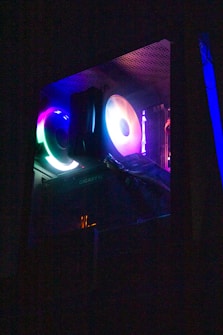 A computer case with vibrant RGB lighting inside, illuminating fans and other components. The colors include shades of blue, purple, and green, creating a colorful glow. The interior is visible through a transparent side panel.