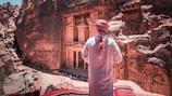 A person wearing traditional attire stands overlooking a historical site carved into rocky cliffs. The architecture displays intricate designs with columns and carvings, and several people are present below, emphasizing the site's monumental scale.