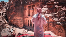 A person wearing traditional attire stands overlooking a historical site carved into rocky cliffs. The architecture displays intricate designs with columns and carvings, and several people are present below, emphasizing the site's monumental scale.