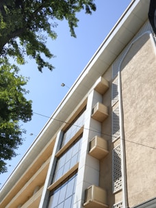 A multi-story building with a modern design featuring large glass windows and decorative elements on the facade. Several air conditioning units are visible attached to the walls. The perspective is from below, looking up at the building against a clear blue sky. Green tree leaves frame the left side, adding a natural element to the urban scene.