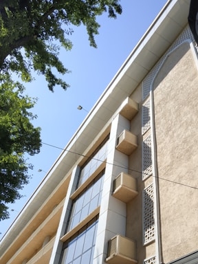 A multi-story building with a modern design featuring large glass windows and decorative elements on the facade. Several air conditioning units are visible attached to the walls. The perspective is from below, looking up at the building against a clear blue sky. Green tree leaves frame the left side, adding a natural element to the urban scene.