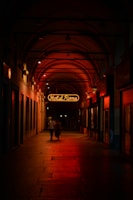 A dimly lit arched corridor with a neon sign reading 'Hotel Roma' glowing in red. Two people walk together under the warm, ambient lighting, which casts reflections on the stone floor. The corridor is lined with closed storefronts.