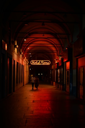 A dimly lit arched corridor with a neon sign reading 'Hotel Roma' glowing in red. Two people walk together under the warm, ambient lighting, which casts reflections on the stone floor. The corridor is lined with closed storefronts.