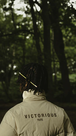 A person with a dark complexion stands in a forest with dense greenery in the background. They are wearing a light-colored jacket with the words 'VICTORIOUS YUTE' printed on the back. Their hair is styled with a yellow stick or accessory.