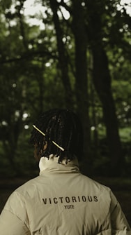 A person with a dark complexion stands in a forest with dense greenery in the background. They are wearing a light-colored jacket with the words 'VICTORIOUS YUTE' printed on the back. Their hair is styled with a yellow stick or accessory.
