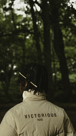 A person with a dark complexion stands in a forest with dense greenery in the background. They are wearing a light-colored jacket with the words 'VICTORIOUS YUTE' printed on the back. Their hair is styled with a yellow stick or accessory.