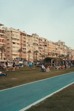 Tall residential buildings with varied facades and balconies line a street. In the foreground, people are sitting, strolling, and lounging on a grass area next to a blue painted pathway. The mood seems relaxed and social.