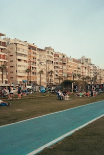 Tall residential buildings with varied facades and balconies line a street. In the foreground, people are sitting, strolling, and lounging on a grass area next to a blue painted pathway. The mood seems relaxed and social.