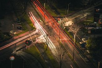 An aerial view of a city intersection at night, marked by streaks of red and white light from moving vehicles. The scene is illuminated by streetlights, showing multiple roads, parked cars, and bare trees alongside the streets. The glowing traffic lights indicate both red and green signals.