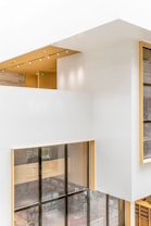 A modern architectural interior with large windows and a clean, minimalist design. The walls are primarily white, interspersed with light wood accents. Natural light floods the space through expansive floor-to-ceiling windows, creating an open and airy atmosphere. The ceiling features embedded linear lighting and a wood-paneled section, adding warmth to the overall aesthetic.