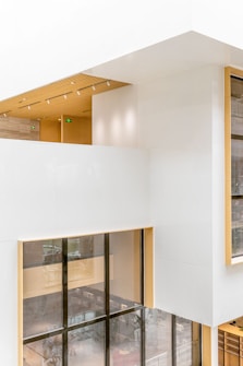 A modern architectural interior with large windows and a clean, minimalist design. The walls are primarily white, interspersed with light wood accents. Natural light floods the space through expansive floor-to-ceiling windows, creating an open and airy atmosphere. The ceiling features embedded linear lighting and a wood-paneled section, adding warmth to the overall aesthetic.