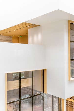 A modern architectural interior with large windows and a clean, minimalist design. The walls are primarily white, interspersed with light wood accents. Natural light floods the space through expansive floor-to-ceiling windows, creating an open and airy atmosphere. The ceiling features embedded linear lighting and a wood-paneled section, adding warmth to the overall aesthetic.