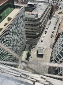 An aerial view of an urban area featuring modern architecture with tall buildings, a cobblestone street lined with parked cars, and several pedestrians walking on the pavement. Greenery can be seen on terraces, and the buildings are distinctly geometric with sharp angles and glass facades.
