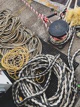 Numerous thick and thin ropes and cords are piled and coiled on a concrete surface. The ropes are of various materials and colors, including yellow and black twists and a reddish spiral. There is a black bollard with ropes tied around it in the top right corner, and parts of the ground feature a metallic surface.
