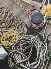 Numerous thick and thin ropes and cords are piled and coiled on a concrete surface. The ropes are of various materials and colors, including yellow and black twists and a reddish spiral. There is a black bollard with ropes tied around it in the top right corner, and parts of the ground feature a metallic surface.