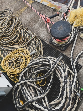 Numerous thick and thin ropes and cords are piled and coiled on a concrete surface. The ropes are of various materials and colors, including yellow and black twists and a reddish spiral. There is a black bollard with ropes tied around it in the top right corner, and parts of the ground feature a metallic surface.
