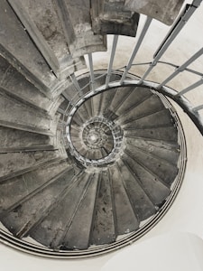 A spiral staircase viewed from above, with metal railings and stone steps winding downward. The perspective creates a dizzying effect as the stairs spiral tightly around a central axis.