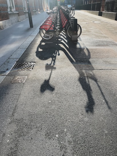 A row of rental bicycles is parked on a city street, casting long shadows across the pavement under bright sunlight. The bikes have red panels with a logo on them, and they are neatly lined up in their docking stations. The surrounding area includes a sidewalk, building facades, and a street light.