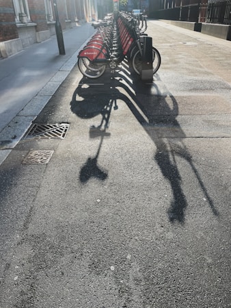 A row of rental bicycles is parked on a city street, casting long shadows across the pavement under bright sunlight. The bikes have red panels with a logo on them, and they are neatly lined up in their docking stations. The surrounding area includes a sidewalk, building facades, and a street light.