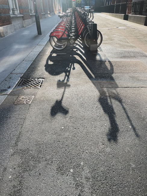 A row of rental bicycles is parked on a city street, casting long shadows across the pavement under bright sunlight. The bikes have red panels with a logo on them, and they are neatly lined up in their docking stations. The surrounding area includes a sidewalk, building facades, and a street light.