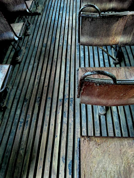 An arrangement of vintage wooden chairs with metal frames, placed on a floor made of long, narrow wooden slats. The chairs appear aged, showing some wear and patina, and the lighting adds a moody, atmospheric effect with subtle shadows.