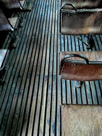 An arrangement of vintage wooden chairs with metal frames, placed on a floor made of long, narrow wooden slats. The chairs appear aged, showing some wear and patina, and the lighting adds a moody, atmospheric effect with subtle shadows.