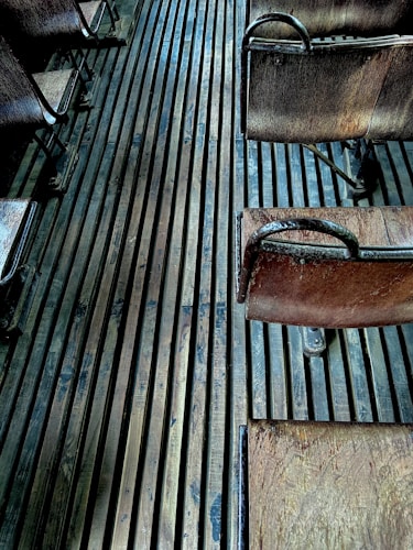 An arrangement of vintage wooden chairs with metal frames, placed on a floor made of long, narrow wooden slats. The chairs appear aged, showing some wear and patina, and the lighting adds a moody, atmospheric effect with subtle shadows.