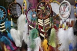 A collection of colorful dreamcatchers, featuring intricate beadwork and feathers in various colors. Some dreamcatchers have circular patterns with a woven web, while others include small paintings or images. The feathers are vibrant, ranging from blue and green to yellow and red.