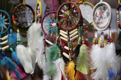 A collection of colorful dreamcatchers, featuring intricate beadwork and feathers in various colors. Some dreamcatchers have circular patterns with a woven web, while others include small paintings or images. The feathers are vibrant, ranging from blue and green to yellow and red.