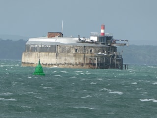 A fortress-like structure situated in a body of water with a green buoy floating nearby. The fortress has a cylindrical design with rugged stone walls and features a red and white striped lighthouse on top. The water is choppy, and the background shows a distant shoreline with trees.
