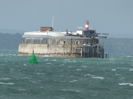 A fortress-like structure situated in a body of water with a green buoy floating nearby. The fortress has a cylindrical design with rugged stone walls and features a red and white striped lighthouse on top. The water is choppy, and the background shows a distant shoreline with trees.