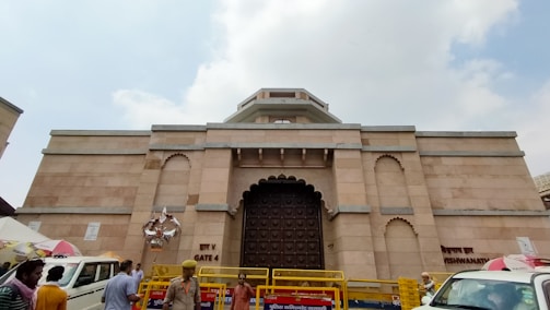 A large stone building with an ornate wooden door and decorative arches. People and vehicles are present in front, including police officers and yellow barricades. The structure features a sign that reads 'Kashi Vishwanath.' The sky is partly cloudy.