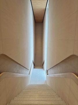 A modern, minimalistic staircase with clean lines and light-colored walls. The view is from the top, looking down a long set of perfectly straight steps. The walls and ceiling create a narrow perspective, enhancing the depth of the image.