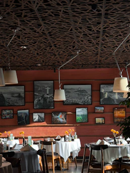 A stylish restaurant interior features several tables adorned with white tablecloths and small vases holding yellow flowers. The wooden walls are decorated with framed photographs and paintings, creating an artistic ambiance. The ceiling has a unique patterned design with multiple hanging lamps providing warm lighting.