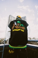 A person wearing a black jacket with a back graphic of a character holding a gun and the words 'Victorious Yute'. The individual is facing away from the camera. They are wearing a yellow cap, standing on a rooftop with a cityscape and a tall building in the background.