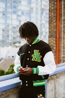 A person with dreadlocks is standing outdoors, looking down at a smartphone. They are wearing a black varsity jacket with white sleeves and colorful trims, featuring large letters on one side. The background includes a blurred urban setting with buildings and a brick wall.