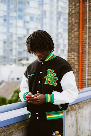 A person with dreadlocks is standing outdoors, looking down at a smartphone. They are wearing a black varsity jacket with white sleeves and colorful trims, featuring large letters on one side. The background includes a blurred urban setting with buildings and a brick wall.